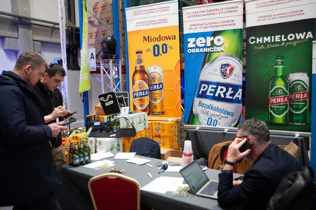 Stoisko z reklamami piwa bezalkoholowego i napoj&oacute;w alkoholowych na targach w Gdańsku, z uczestnikami rozmawiającymi przy stole.