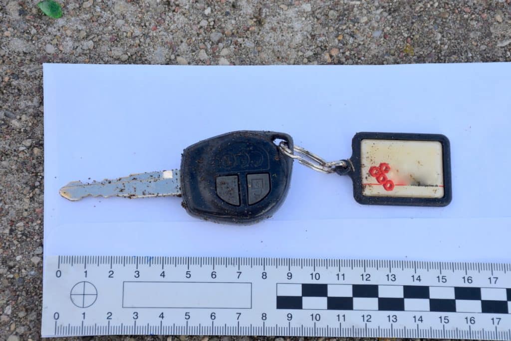 Zdjęcie kluczyka samochodowego z tabliczką, leżące na ziemi w lesie, obok linijki do pomiaru.