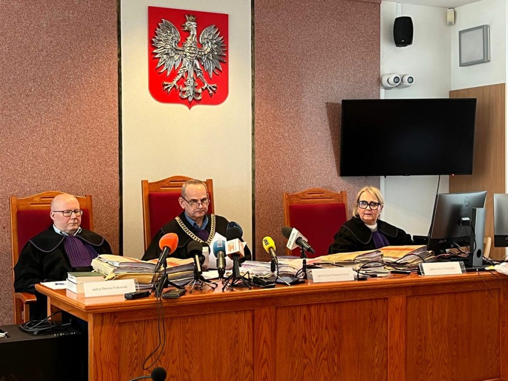 Sędziowie podczas rozprawy w sali sądowej z orłem na ścianie za nimi.