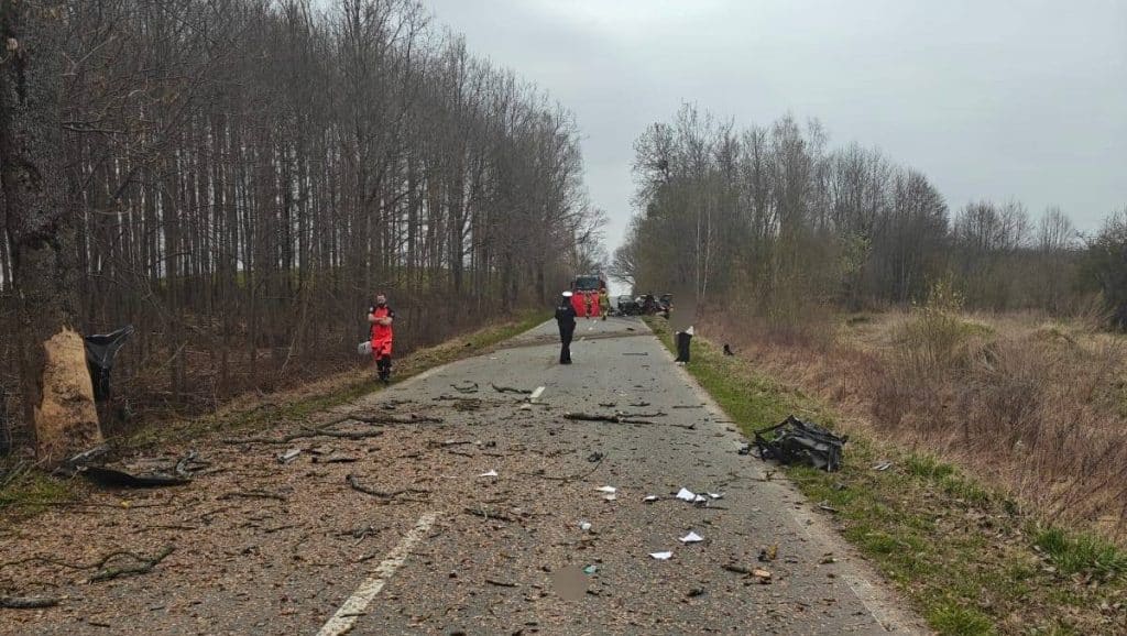 Zniszczony samoch&oacute;d i rozbitych elementy na drodze po wypadku, w tle służby ratunkowe.