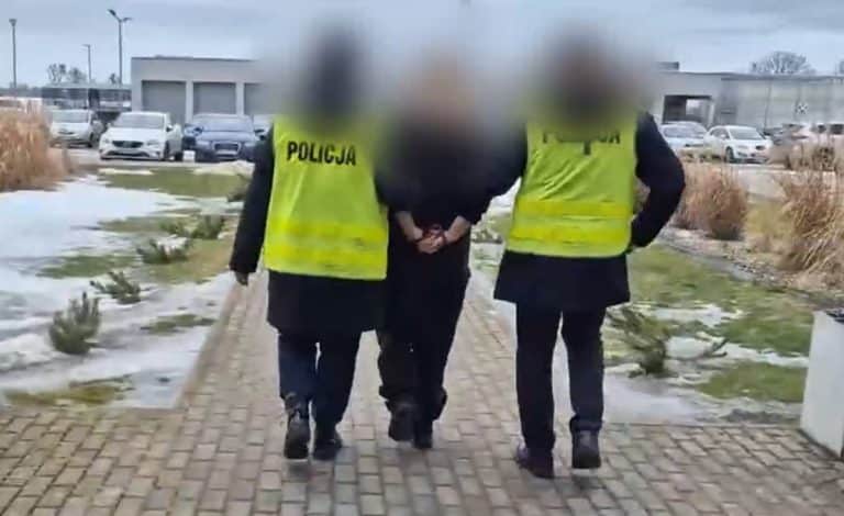 Policjanci prowadzą zatrzymaną kobietę w kamizelkach policyjnych na zewnątrz budynku.