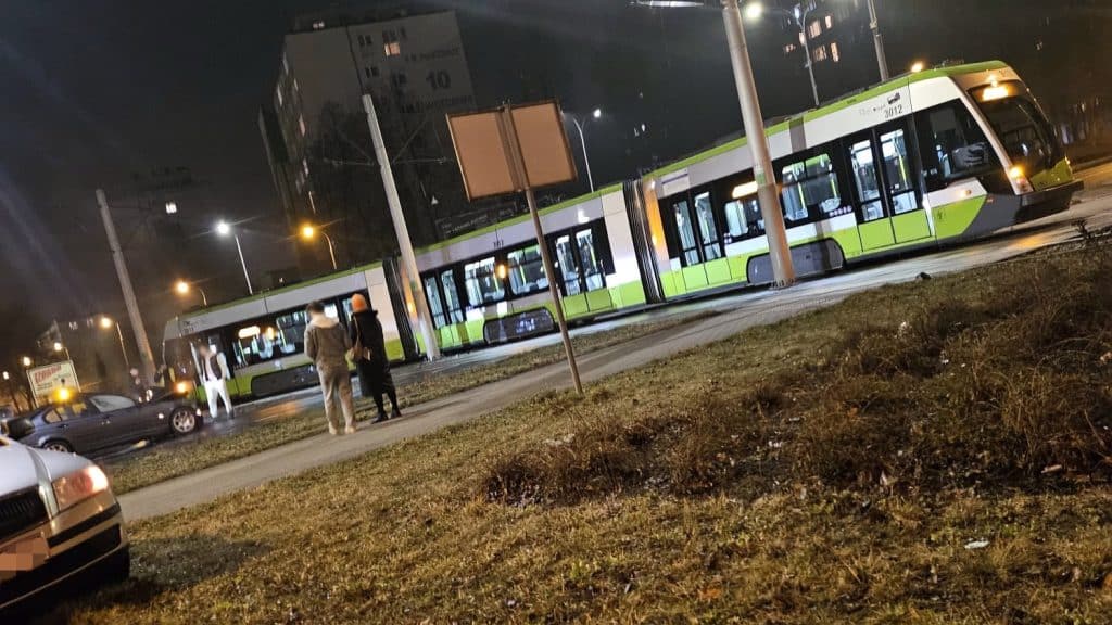 Kolizja samochodu z tramwajem na rondzie w Olsztynie, kilka os&oacute;b na chodniku, noc, pojazdy i tramwaj na miejscu.