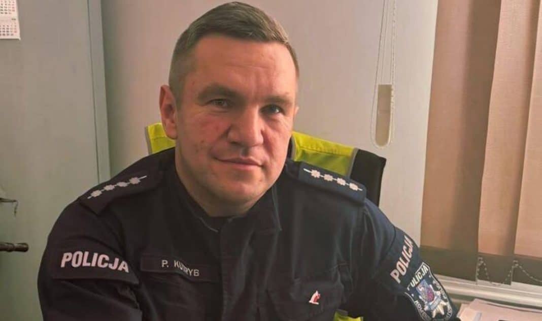 Policjant w mundurze siedzi w pomieszczeniu, za nim widoczny jest telefon i zasłony.