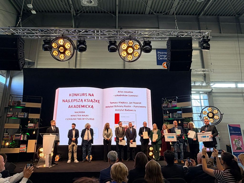 Na scenie podczas wręczenia nagr&oacute;d, grupa os&oacute;b trzyma certyfikaty i trofea, w tle ekran z informacją o konkursie.