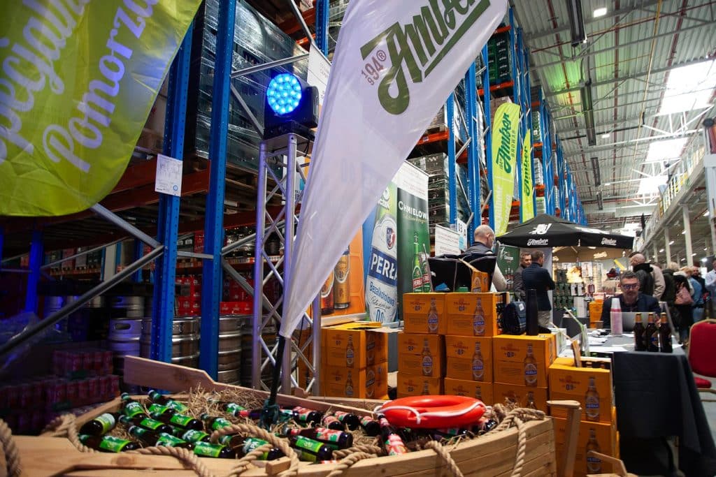 Stoisko targowe z produktami alkoholowymi i napojami, otoczone flagami i uczestnikami wydarzenia.