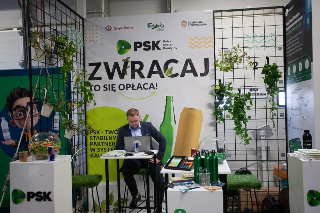 Stoisko targowe z logo PSK, roślinami i materiałami promocyjnymi, na tle siatki i baner&oacute;w.