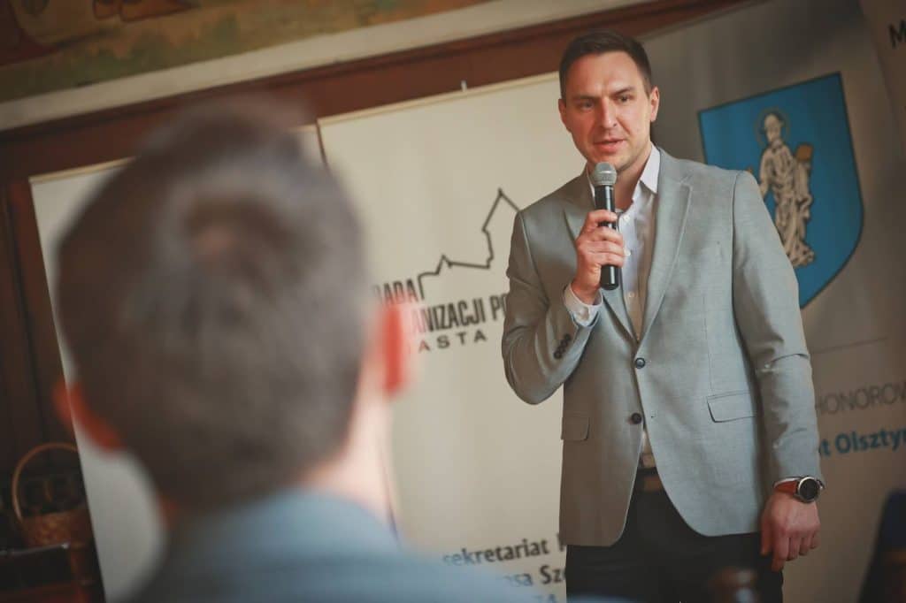 Mężczyzna w garniturze przemawia do zgromadzonych podczas spotkania organizacji społecznych w Olsztynie.