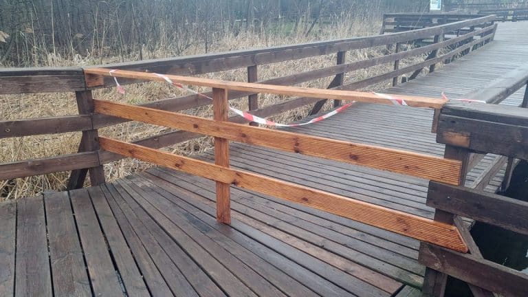 Fragment drewnianego molo nad jeziorem, zniszczona część i barierka, na tle natury.