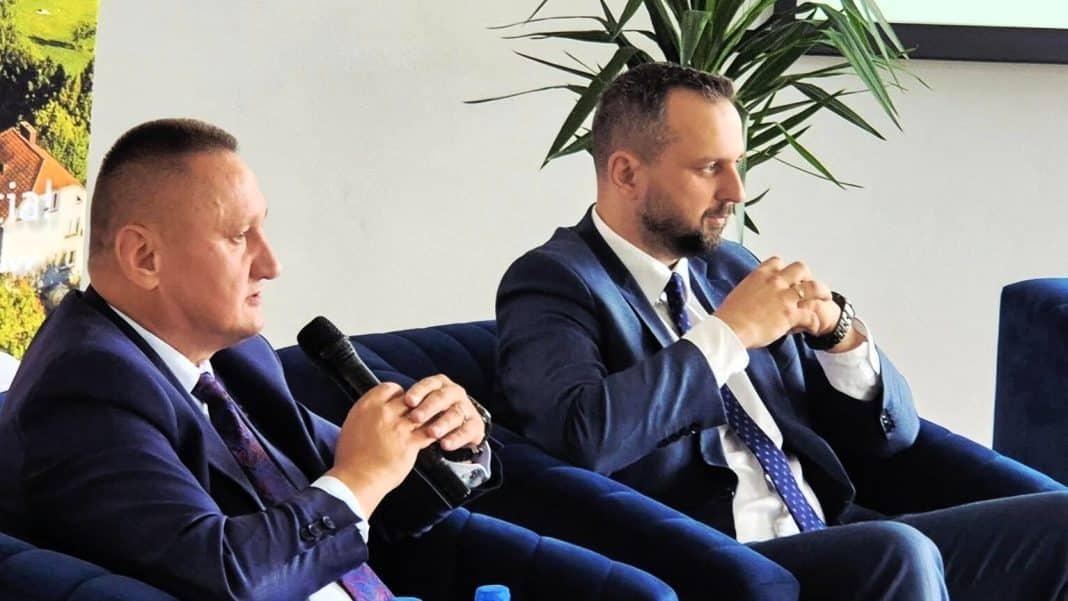 Dw&oacute;ch mężczyzn w garniturach podczas konferencji, jeden z nich trzyma mikrofon.