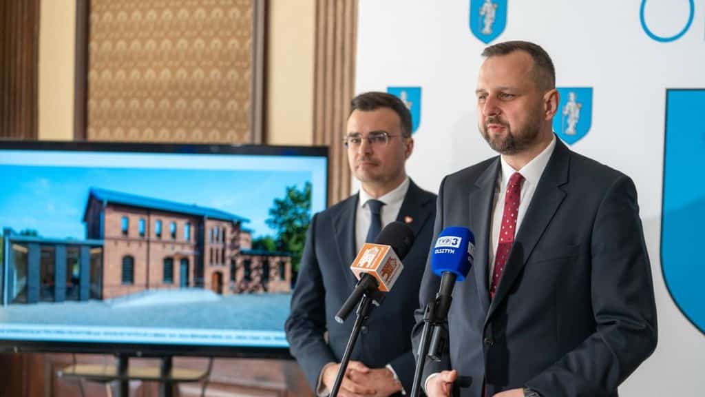 Dw&oacute;ch mężczyzn w garniturach podczas konferencji prasowej, obok ekran z wizualizacją odrestaurowanego zabytku.