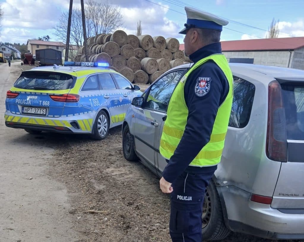 Policjant zatrzymuje samoch&oacute;d na drodze, w tle widoczne policyjne radiowozy i wiejskie otoczenie.