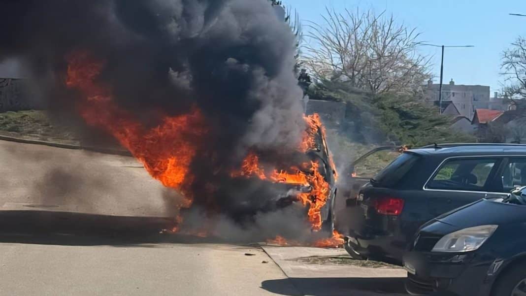 Samoch&oacute;d marki BMW w płomieniach na ulicy, z dymem unoszącym się nad pojazdem.