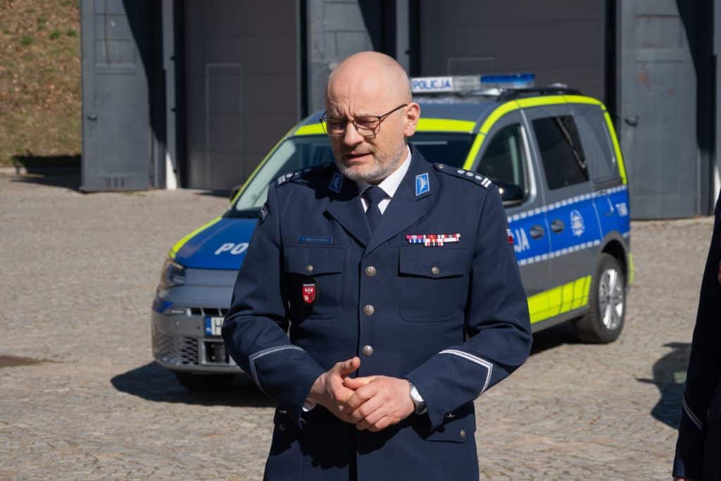 Policjant w mundurze stoi przed nowym radiowozem na Mazurach, w tle policyjny samoch&oacute;d w barwach policyjnych.