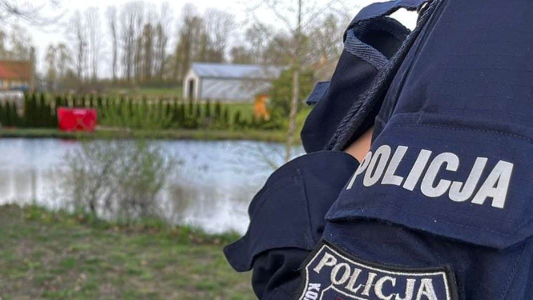 Policjant przy stawie na Mazurach, w tle widoczny wodny akwen i domy.