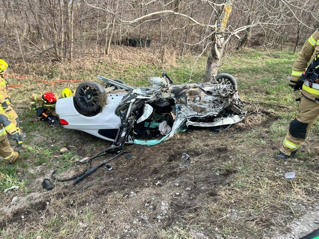 Rozbity samoch&oacute;d leży na poboczu po karambolu na Mazurach, wok&oacute;ł strażacy w akcji ratunkowej.