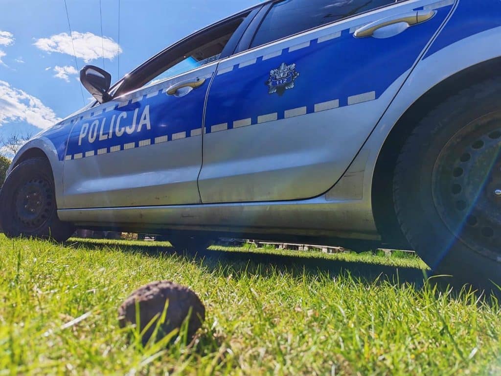 Policja na miejscu zdarzenia w ogrodzie, widok na samoch&oacute;d policyjny i ziemię z wykopanym przedmiotem.