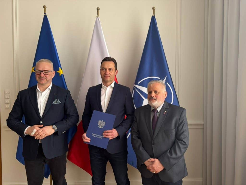 Trzej mężczyźni stojący przed flagami UE, Polski i NATO podczas wręczenia powołań.