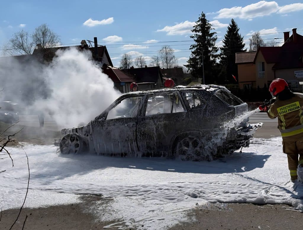 Strażacy gaszą spalony samoch&oacute;d na ulicy w Elblągu, wok&oacute;ł widoczne domy i niebo z chmurami.