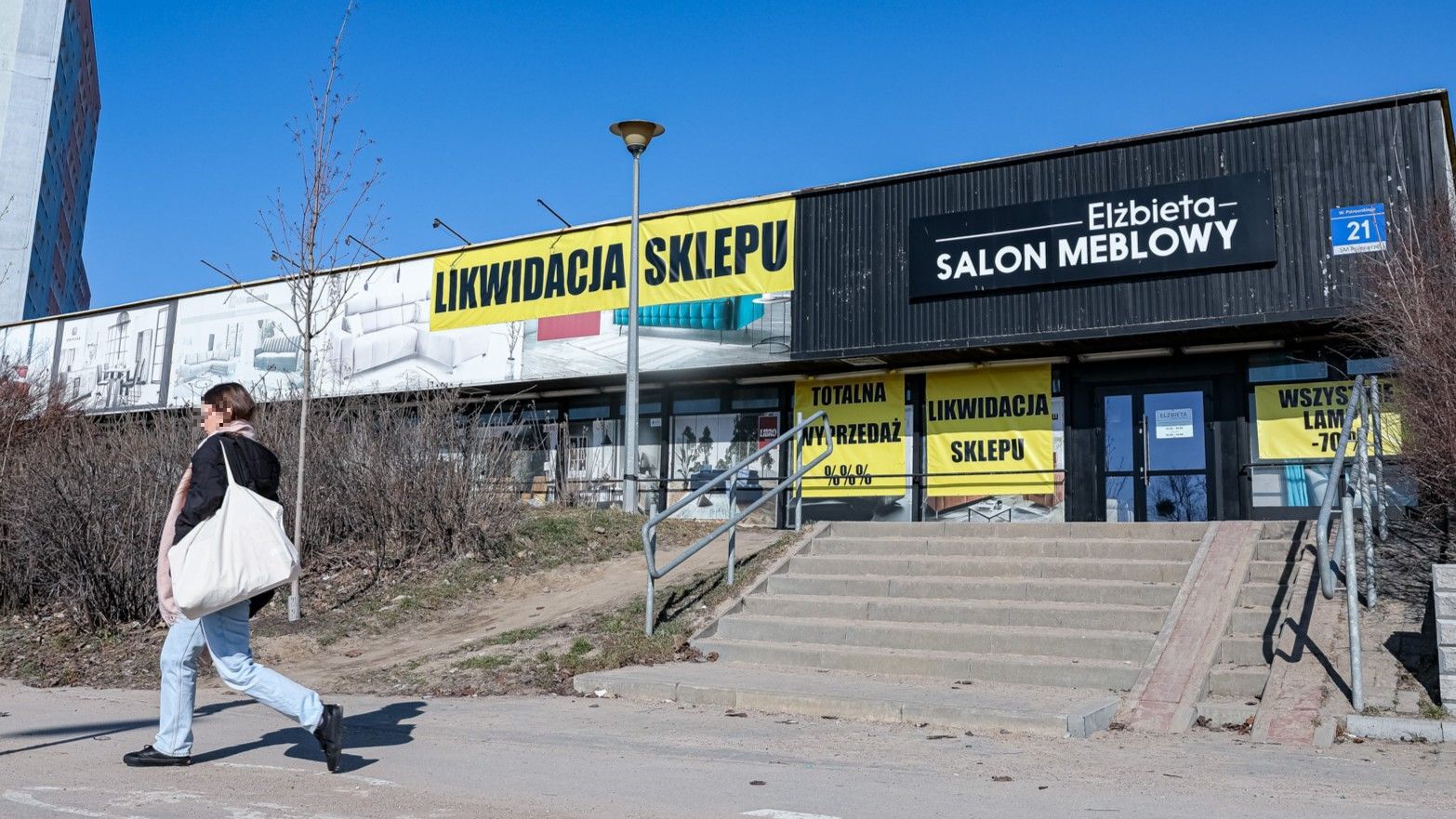 Widok z zewnątrz na sklep meblowy z dużymi plakatami wyprzedaży i likwidacji, na tle nieba.
