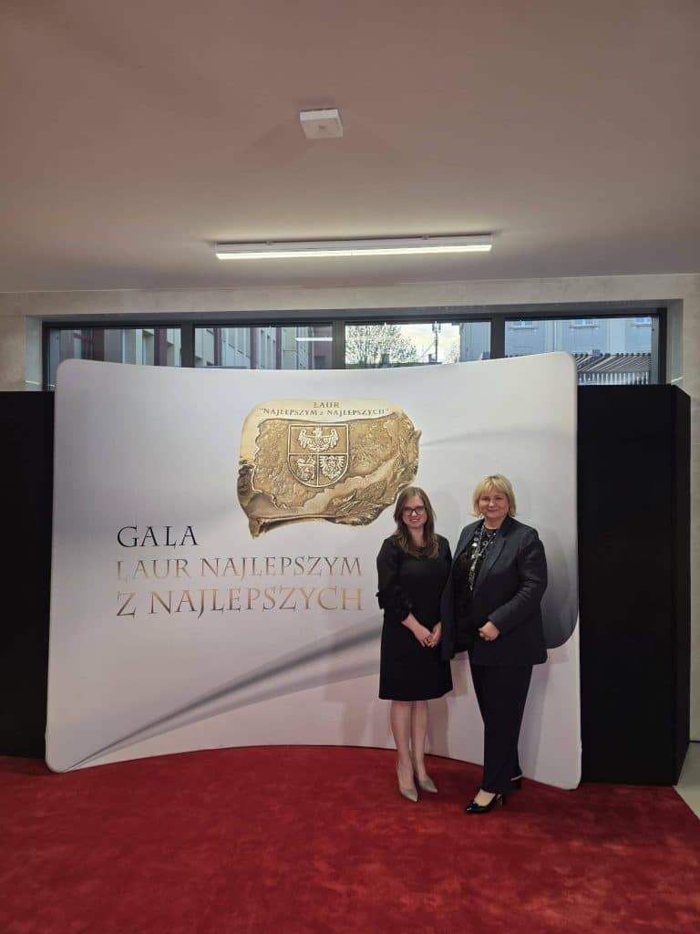 Dwie kobiety stoją na tle dużego banera z napisem 'Gala Laur Najlepszym z Najlepszych'.