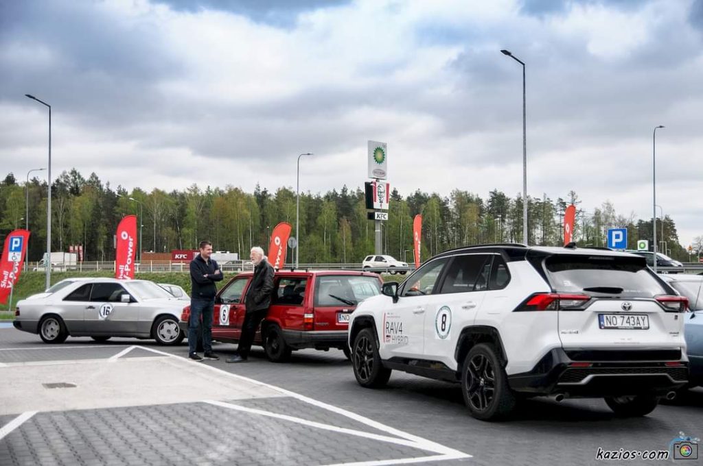 Uczestnicy wydarzenia rozmawiają przy samochodach na parkingu, w tle stacje benzynowe i reklamy.