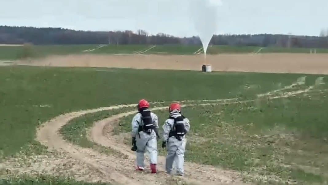 Dwoje ratownik&oacute;w w kombinezonach i kaskach na polu, w tle wystrzał z urządzenia na tle nieba.