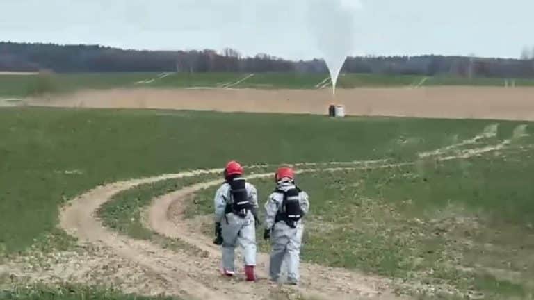 Dwoje ratownik&oacute;w w kombinezonach i kaskach na polu, w tle wystrzał z urządzenia na tle nieba.