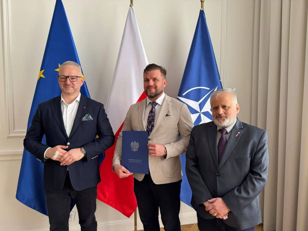 Trzech mężczyzn w formalnych ubraniach stoi przed flagami UE, Polski i NATO podczas uroczystości wręczenia powołań.