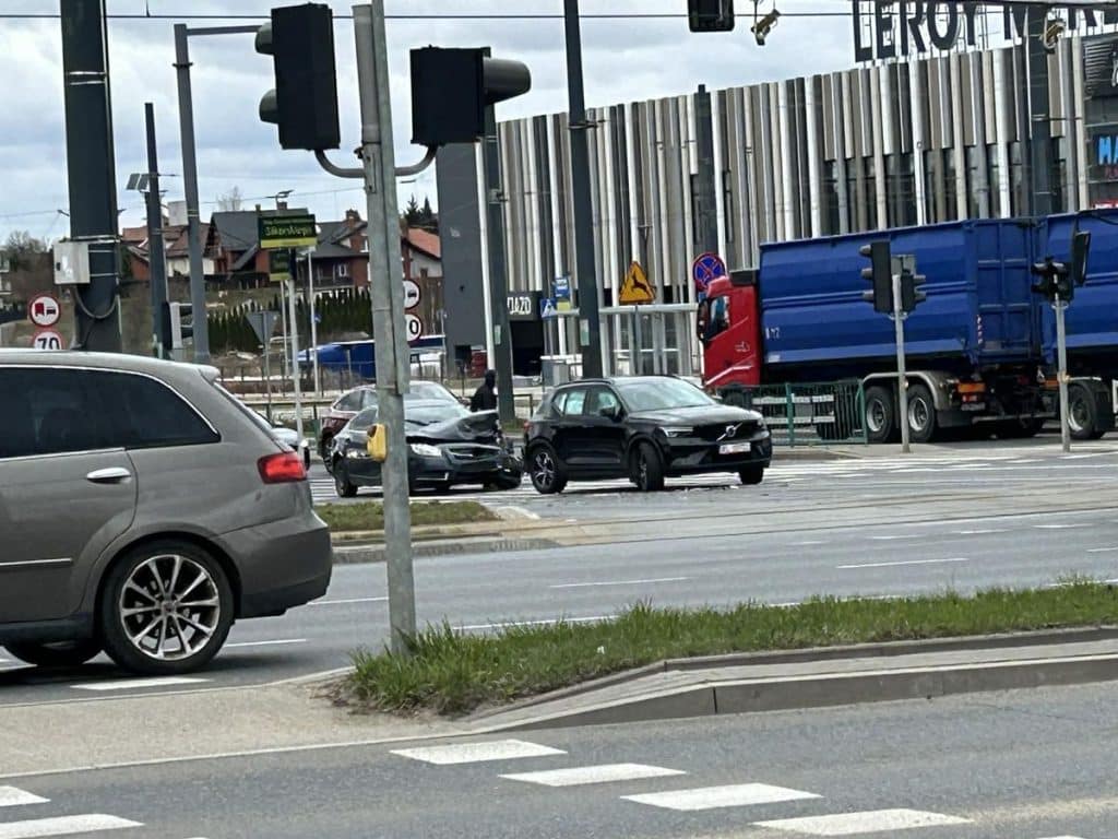Zdjęcie z miejsca kolizji na skrzyżowaniu przy Galerii Warmińskiej, z uszkodzonymi samochodami i słupkami.