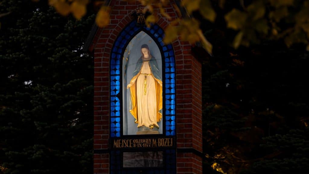 Oświetlona figura Matki Bożej w kaplicy, otoczona ciemnym lasem, z widocznymi złotymi gwiazdami na niebie.