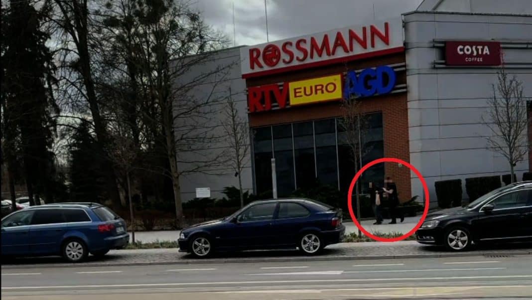 Zdjęcie przedstawia grupę os&oacute;b na chodniku przy centrum handlowym Aura w Olsztynie, z zaznaczonym czerwonym k&oacute;łkiem.