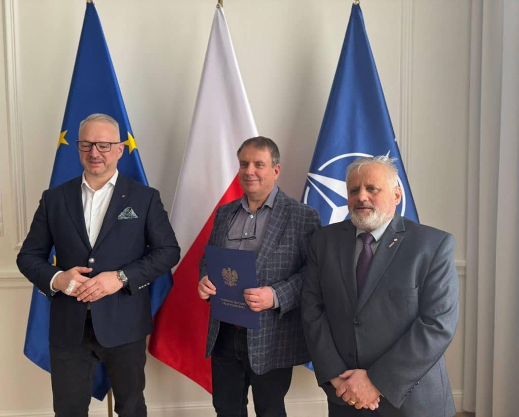 Trzej mężczyźni stojący przed flagami UE, Polski i NATO podczas wręczenia powołań.