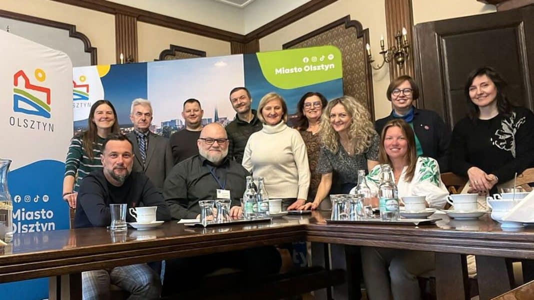 Grupa ludzi podczas spotkania w sali konferencyjnej z banerami Miasta Olsztyn w tle.