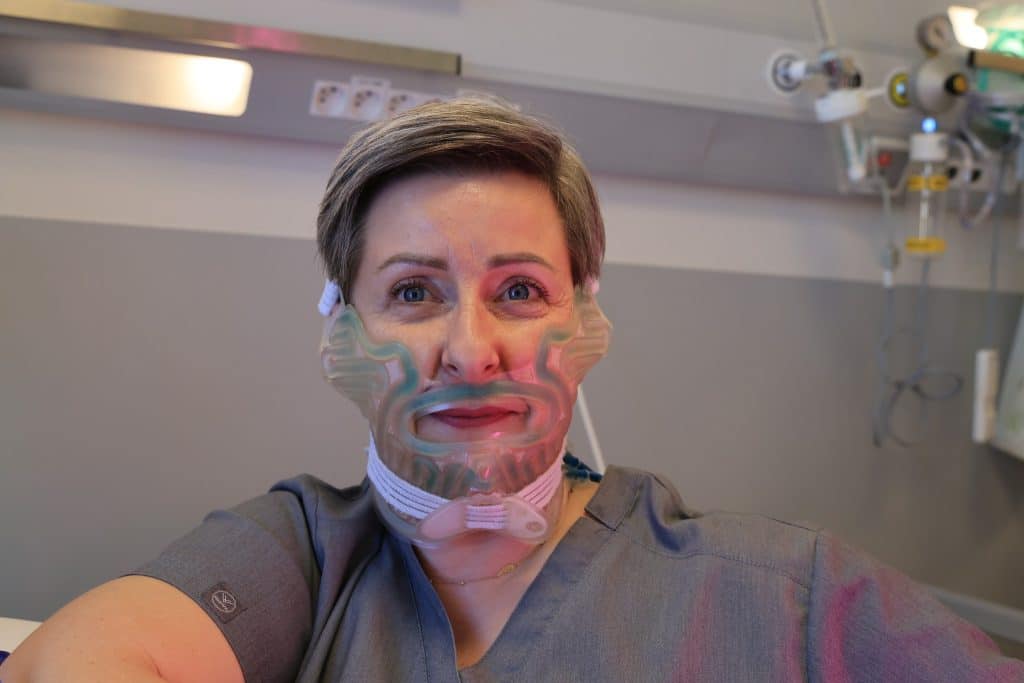 Kobieta w szpitalu z maską chłodzącą na twarzy, w tle urządzenia medyczne.