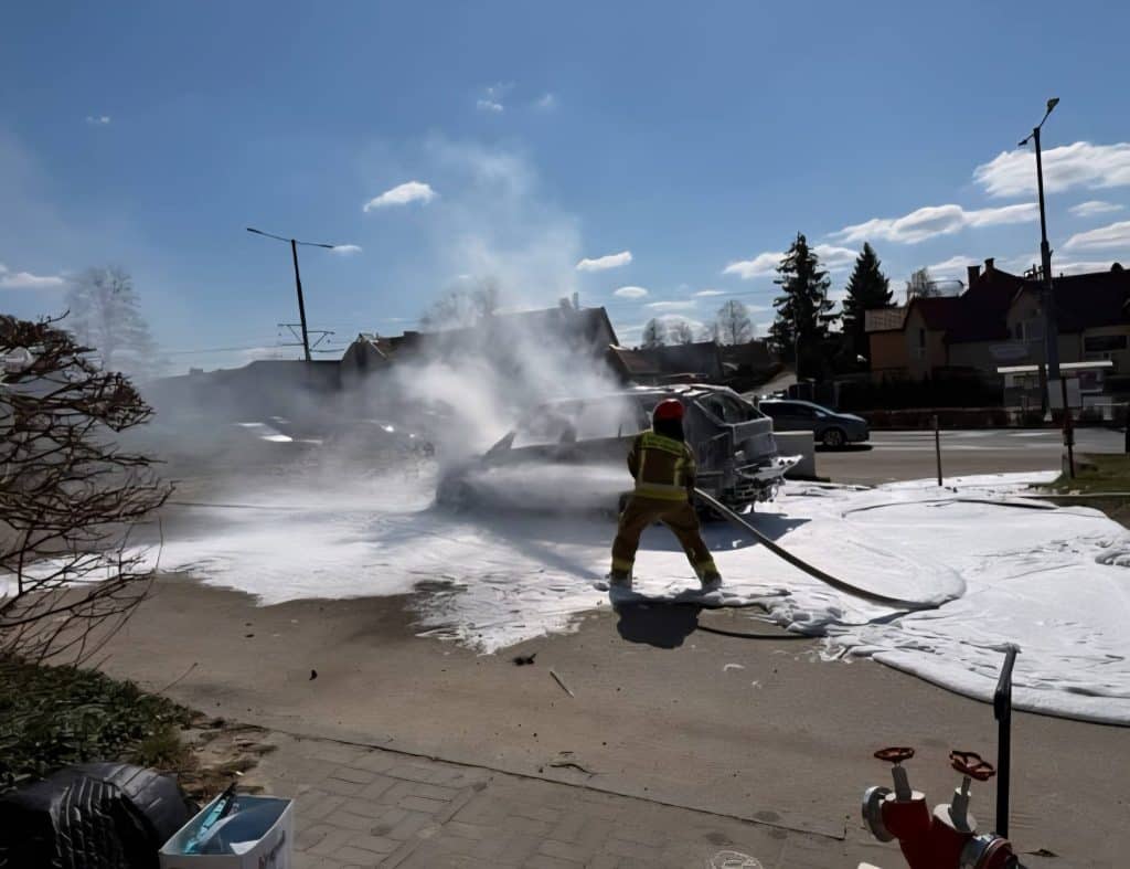 Strażak gaszący pożar samochodu na ulicy, unoszą się kłęby dymu, w tle domy i niebo.