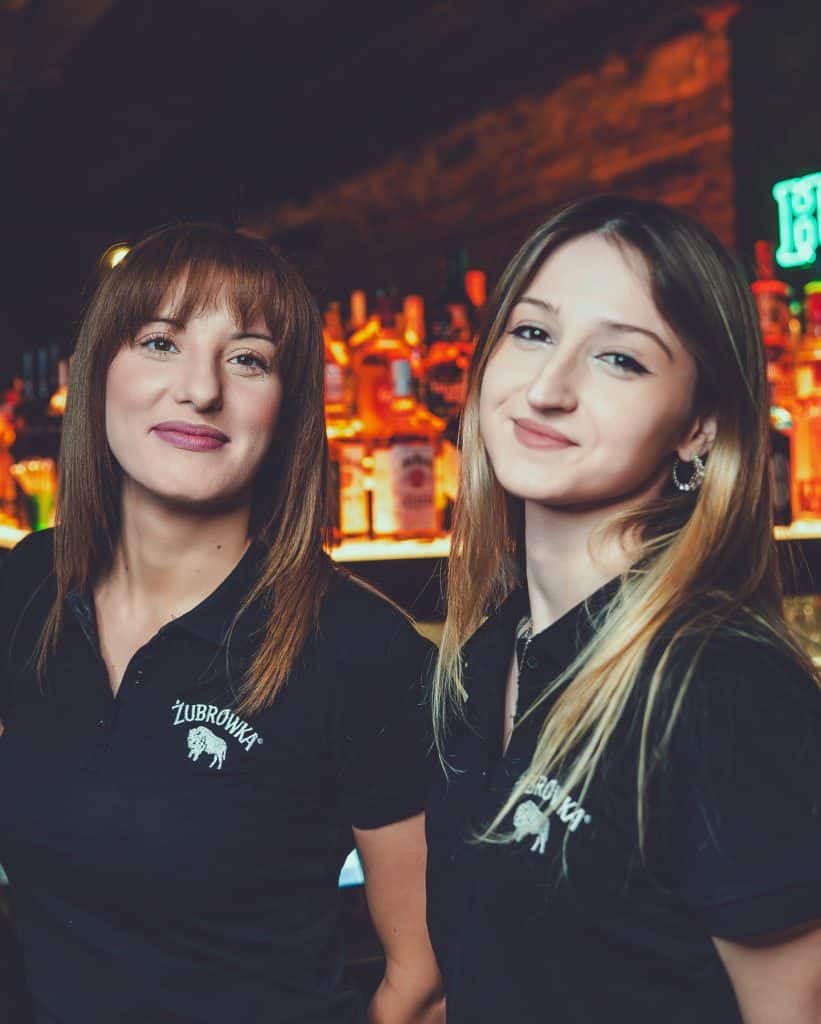 Dwie kobiety w klubie nocnym przy barze, oświetlone neonami, uśmiechnięte, w czarnych koszulkach z logo.