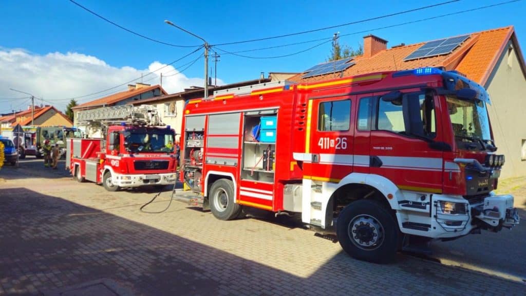 Strażacy w akcji przed budynkiem, w tle dom z panelami słonecznymi na dachu.