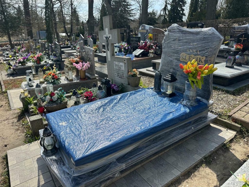 Nagrobek przykryty folią na cmentarzu, wok&oacute;ł kwiaty i znicze, w tle inne groby.