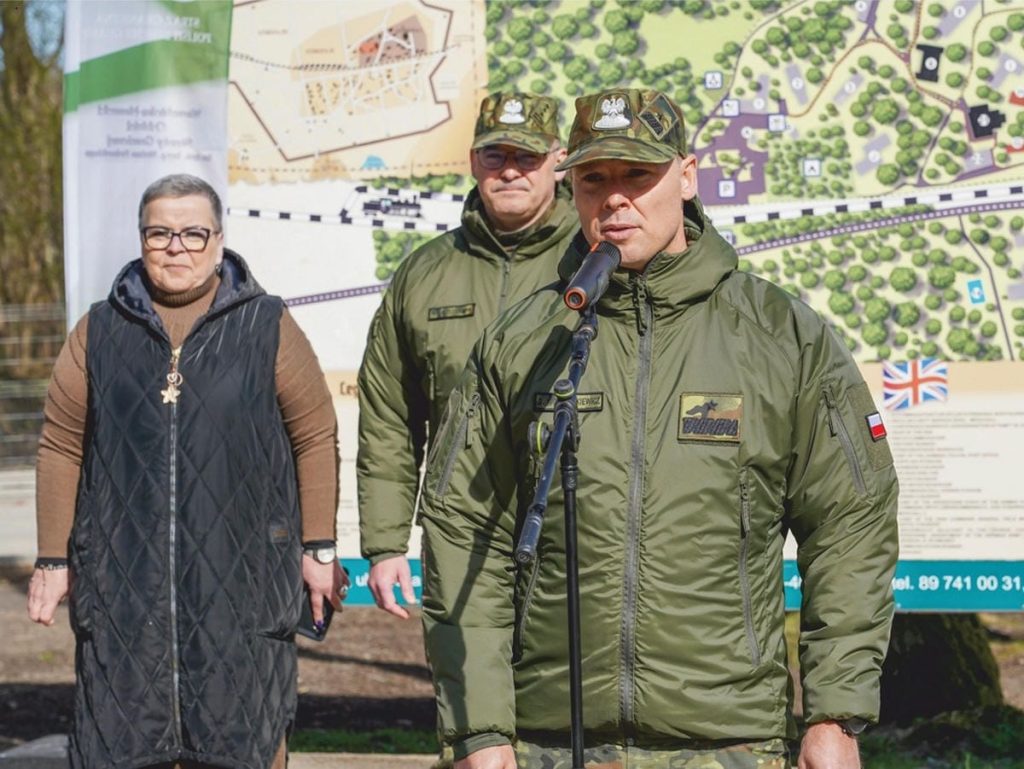 Grupa os&oacute;b podczas wydarzenia sadzenia drzew w Wilczym Szańcu na Mazurach, z mapą w tle.
