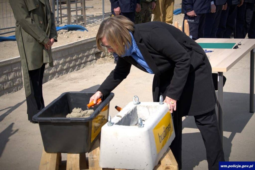 Kobieta w czarnym płaszczu kopie beton w trakcie ceremonii rozpoczęcia budowy nowej komendy policji na Mazurach.