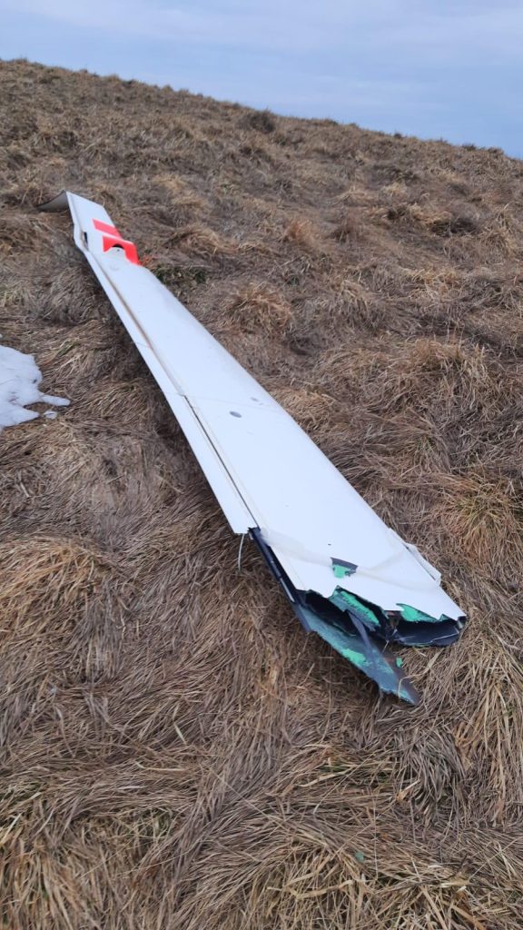 Złamany fragment szybowca leży na trawiastej powierzchni w g&oacute;rach, w tle niebo.