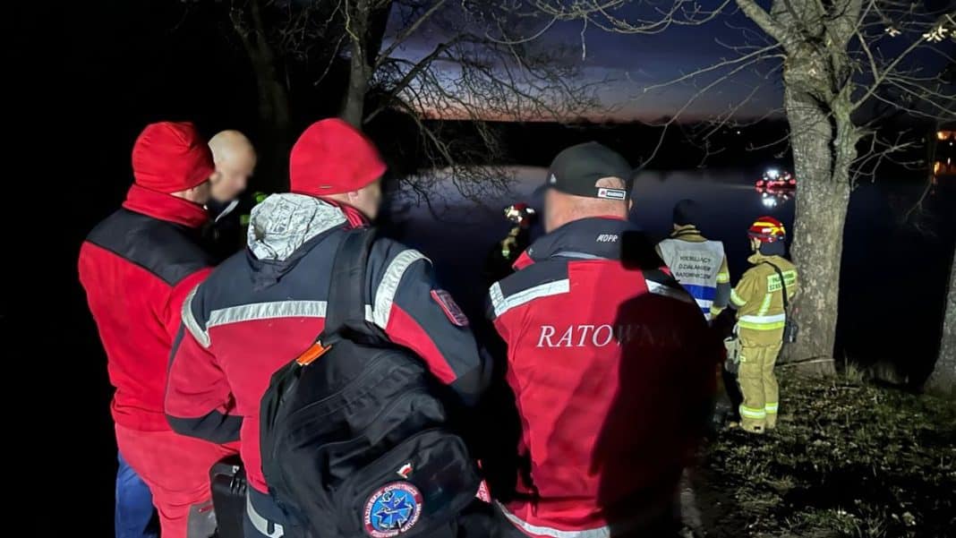 Grupa ratownik&oacute;w i służb poszukiwawczych przy jeziorze nocą, w tle widoczny wodny krajobraz.