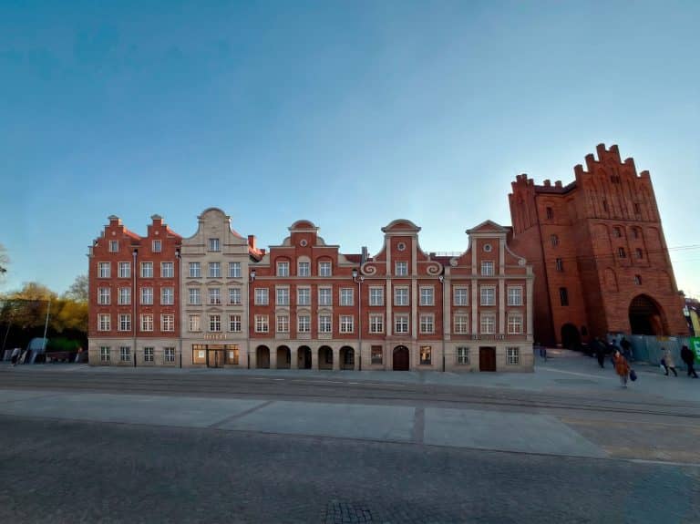 Widok na zabytkowe budynki przy Wysokiej Bramie w Olsztynie, z wyraźnym niebem i ulicą na pierwszym planie.