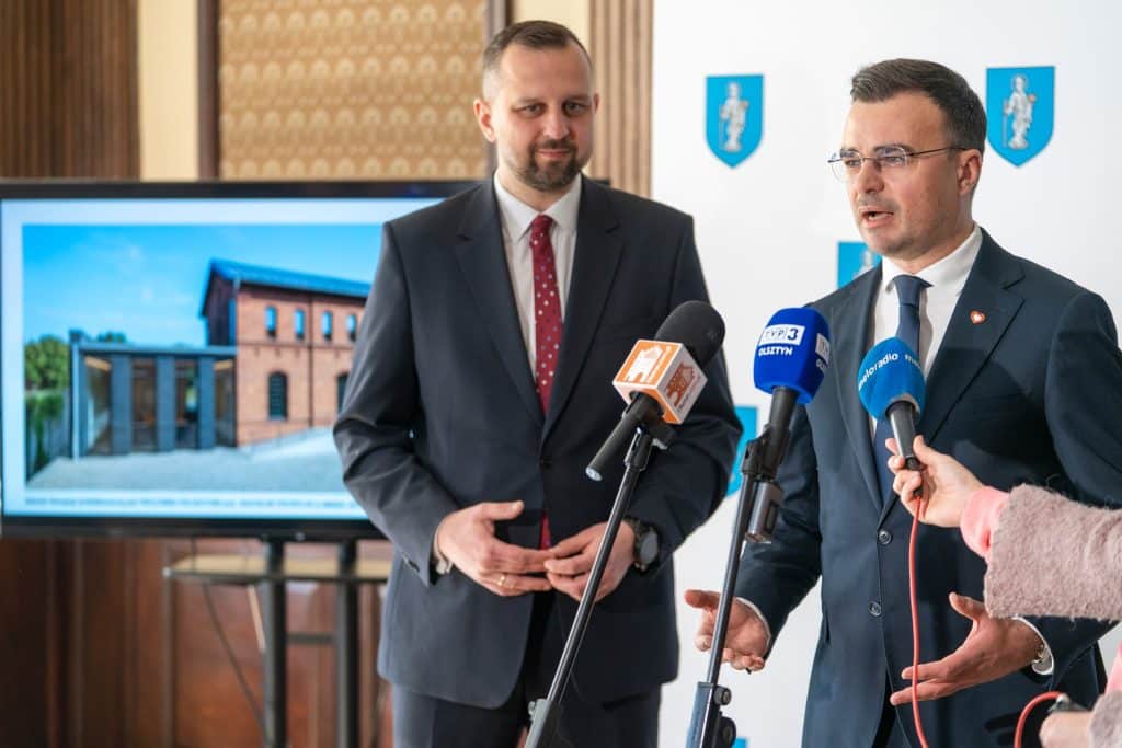 Dw&oacute;ch mężczyzn w garniturach podczas konferencji prasowej, za nimi ekran z wizualizacją obiektu.