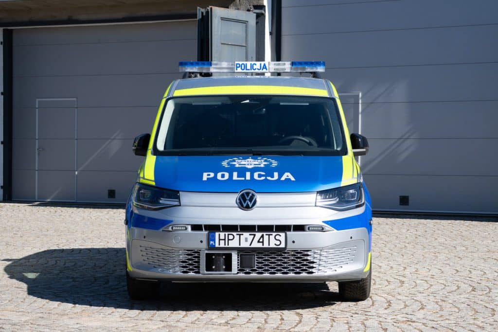 Nowy radiow&oacute;z policyjny na Mazurach, w barwach niebiesko-ż&oacute;łtych, zaparkowany na placu przed budynkiem.