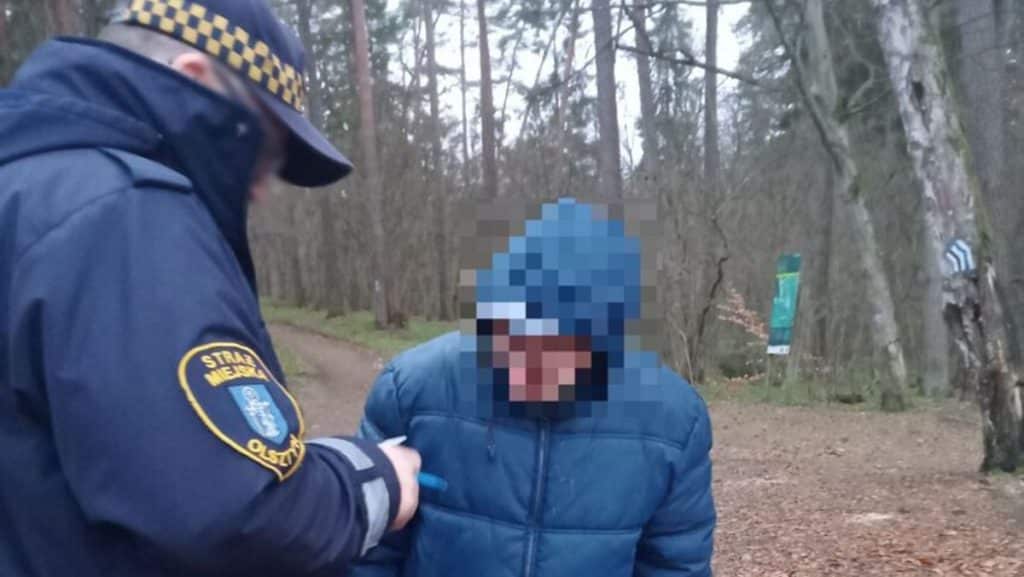 Mężczyzna w kapturze rozmawia z funkcjonariuszem na tle lasu, zachowując się nietypowo.