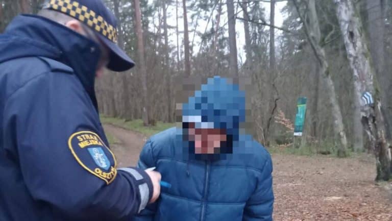 Mężczyzna w kapturze rozmawia z funkcjonariuszem na tle lasu, zachowując się nietypowo.