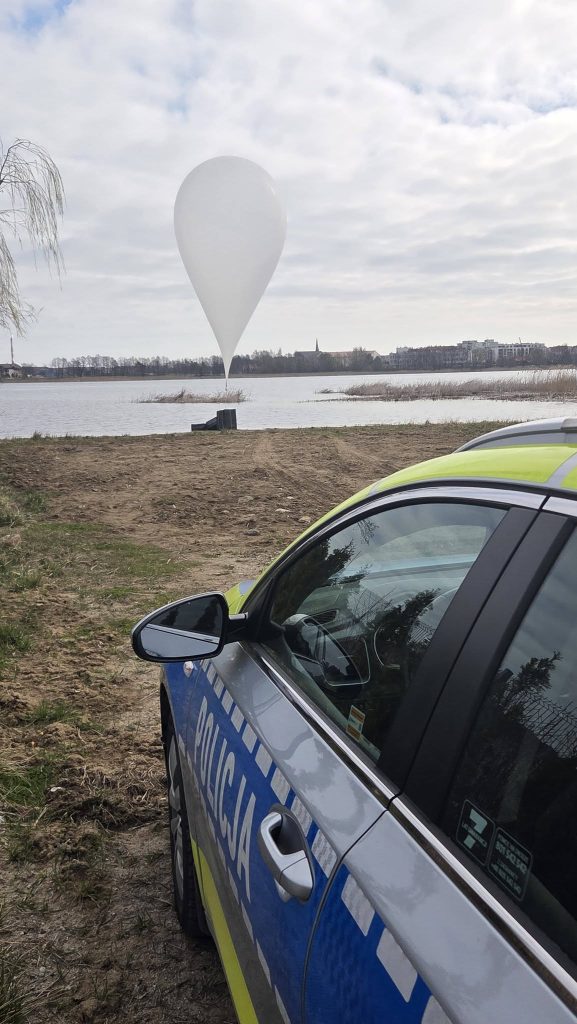 Policja na brzegu jeziora, w tle balon unoszący się nad wodą, w okolicy pola i drzew.