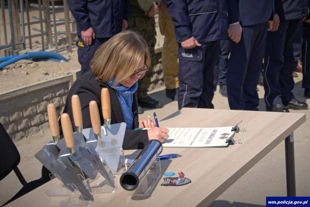 Kobieta podpisuje dokumenty przy stole na placu budowy nowej komendy policji, w tle policjanci.