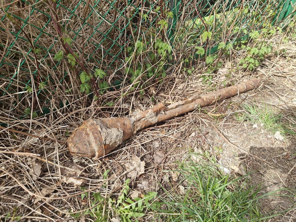 Zardzewiały, długi przedmiot leży na ziemi wśr&oacute;d trawy i krzew&oacute;w, zabezpieczony przez służby nad jeziorem.